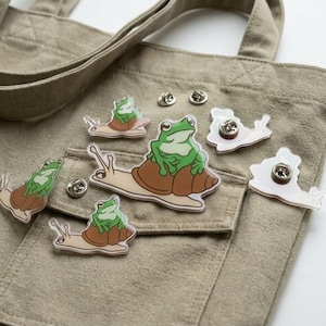 May include: A collection of acrylic pins featuring a green frog sitting on a brown snail shell. The pins are arranged on a beige canvas tote bag. Each pin shows a cartoon-style frog with large eyes resting on the back of a light-colored snail.