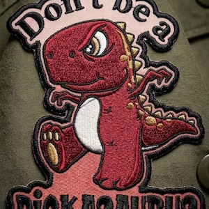 May include: Embroidered patch featuring a cartoon dinosaur with a stern expression. The patch reads "Don't be a Dickasaurus" in black lettering with a pink and black border. The dinosaur is red with gold accents.