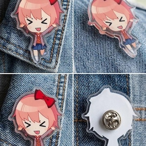 May include: An acrylic pin featuring a chibi-style character with pink hair, a red bow, a grey cardigan, and a blue skirt. The character has a joyful expression with arms raised. The back of the pin shows a silver metal butterfly clutch fastener.
