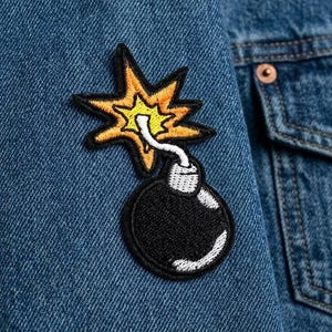 May include: A black and yellow embroidered patch in the shape of a bomb with a starburst design. The patch is sewn onto a blue denim jacket. The bomb has a white fuse and a black body.
