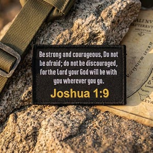 May include: A black patch with white text quoting Joshua 1:9, in gold lettering. The patch is on a rock surface, with a green strap and a map in the background. The text reads: "Be strong and courageous..."