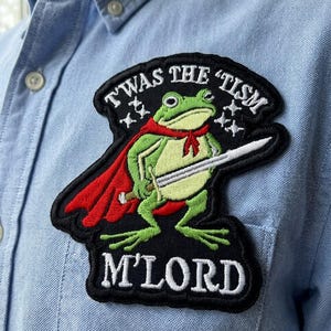 May include: Embroidered patch featuring a green frog in a red cape and holding a sword. The patch has the text "TWAS THE 'TISM M'LORD" in white lettering on a black background. The patch is sewn onto a light blue button-down shirt.