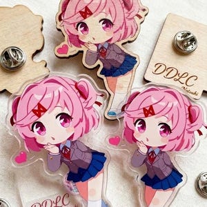 May include: Three pins featuring a pink-haired anime character in a school uniform with a blue skirt and grey blazer. Two pins are clear acrylic with the text DDLC Natsuki, and one is wood-backed. Each pin shows the character blowing a kiss.