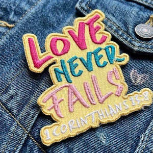 May include: Embroidered patch with the words "LOVE NEVER FAILS" in pink, teal, and yellow. The patch is on a denim jacket. The text "1 CORINTHIANS 13:8" is also included.