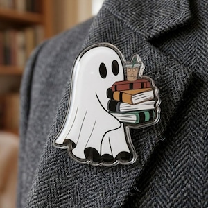 May include: A clear acrylic pin featuring a white ghost with black eyes holding a stack of four books in shades of red, tan, black, and green, topped with a small iced coffee drink with a green straw, attached to a grey herringbone fabric.