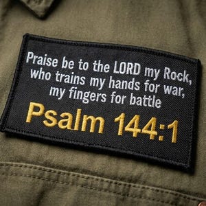 May include: A black rectangular patch with white and gold text, stitched onto an olive green garment. The text reads "Praise be to the LORD my Rock, who trains my hands for war, my fingers for battle Psalm 144:1."