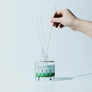 May include: A clear glass diffuser bottle with white reeds. The bottle has a label with the text "Timeless Joy Over Tea" and a graphic of people. A hand is holding a reed.