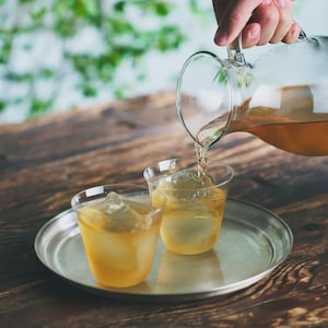 May include: A clear glass jug pouring a golden liquid into two glasses filled with ice cubes. The glasses sit on a silver tray, with a wooden table and blurred green foliage in the background. The jug has a handle.