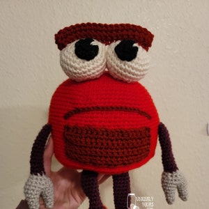 Puede incluir: Un juguete robot de crochet rojo y marrón con ojos blancos y pies naranjas. El robot tiene una expresión triste.