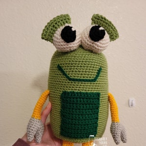 Puede incluir: Juguete de peluche verde tejido a crochet con ojos grandes, una boca sonriente y un cuadrado verde en el pecho. El juguete tiene brazos y piernas amarillos con pies rojos.