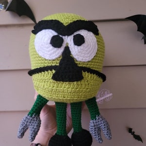 Puede incluir: Un monstruo verde de crochet con ojos negros, nariz negra y cejas negras. El monstruo tiene piernas verdes con pies negros y garras grises.
