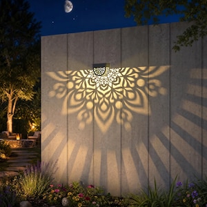 May include: A decorative outdoor wall light casts a floral pattern on a concrete wall. The light fixture is mounted on the wall and emits a warm glow, creating a beautiful display. The scene is set at night with a moon in the sky.