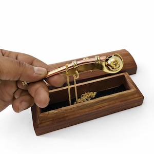 Op de afbeelding: Een goudkleurige fluit met koperen accenten, rustend op een houten sieradendoosje. Een gouden ketting is zichtbaar in de doos. De fluit heeft een decoratief ontwerp aan het uiteinde. De doos is open en onthult een zwart interieur.