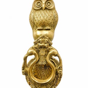 Op de afbeelding: Een koperen deurklopper met een gedetailleerde uil aan de bovenkant, geplaatst boven een centrale figuur met een gezicht en een versierde hoofdtooi. Een ronde ring wordt vastgehouden door twee vogelachtige figuren aan de zijkanten, op een ovaal.