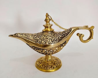 Könnte beinhalten: Eine goldfarbene Dschinn-Lampe mit aufwendigen Blumen- und Rankenmustern. Die Lampe hat einen gebogenen Ausguss, einen geschwungenen Griff und einen kleinen Deckel, der mit einer Kette befestigt ist. Sie steht auf einem runden, verzierten Sockel.