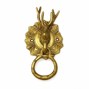 Könnte beinhalten: Ein goldfarbener Türklopfer aus Metall mit einem skulptierten Hirschkopf samt Geweih, der auf einer runden, floral gemusterten Platte montiert ist. Ein runder, segmentierter Ring hängt am Kinn des Hirsches zum Klopfen an der Tür.