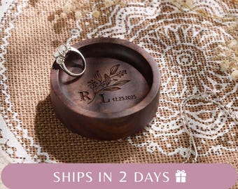 May include: A round dark brown wooden ring dish featuring an engraved floral branch design, the letters R and L, and the date 12.25.2025. A silver ring with a clear stone rests on the edge of the dish, and a silver chain lies nearby.