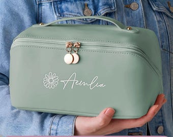 May include: A sage green rectangular toiletry bag with a top handle and a zipper closure featuring two round rose gold pulls. The front displays a white daisy icon and the name Amelia in a cursive script font, set against a smooth matte finish.