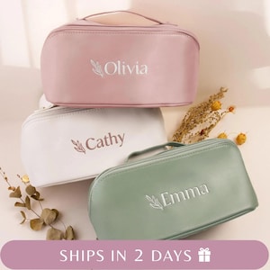 May include: Three personalised cosmetic bags in pink, white, and green. Each bag has a handle and a zip closure. The names "Olivia", "Cathy", and "Emma" are embroidered on the bags in white script with a leaf design.