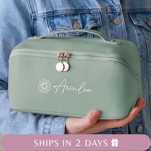 May include: A light green cosmetic bag with a handle and a zipper closure. The bag has gold-colored zipper pulls and a daisy graphic with the name "Amelia" in white script. The bag is held in front of a denim jacket.