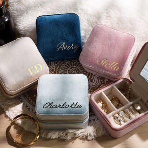 May include: A collection of personalized velvet jewelry boxes in various colors: cream, blue, and pink. Each box is square and features a gold zipper and a name or initials in gold script. One box is open, revealing rings and jewelry.