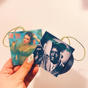 May include: Two miniature vinyl record ornaments. One features a woman in a blue-toned image with the text "THE LIFE OF A". The other has a black and white image of a man with the text "CHROMAKOPIA". Both have gold wire hangers.