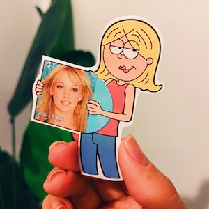 May include: A cartoon sticker of a character holding a photo of a blonde woman. The sticker features a character with blonde hair, wearing a pink top and blue trousers. The photo in the character's hands has the text "Hilary Duff".