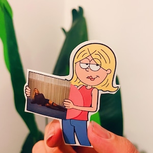 Puede incluir: Una pegatina troquelada con un personaje de dibujos animados sosteniendo una imagen. El personaje de dibujos animados tiene el pelo rubio, una camiseta rosa y vaqueros azules. La imagen muestra a una persona con un atuendo negro. La pegatina está sujeta por una mano.