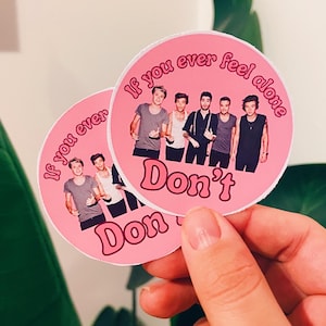 Może przedstawiać: Dwie różowe okrągłe naklejki ze zdjęciem pięciu mężczyzn i napisem "If you ever feel alone Don't". Naklejki są trzymane przez dłoń na tle zielonego, liściastego tła.