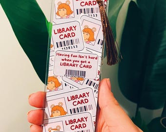 Puede incluir: Un marcapáginas rectangular de plástico transparente con un patrón repetitivo de tarjetas de biblioteca con la ilustración de un personaje de dibujos animados de pelo rubio. El texto dice LIBRARY CARD y Having fun isn't hard when you got a LIBRARY CARD.