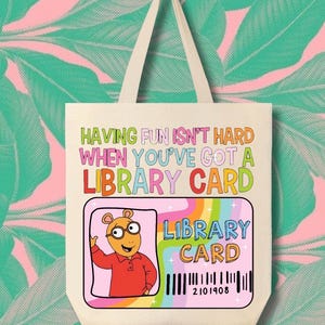 Peut inclure: Sac fourre-tout crème avec le texte "HAVING FUN ISN'T HARD WHEN YOU'VE GOT A LIBRARY CARD." Il présente un design de carte de bibliothèque colorée avec un personnage de dessin animé et un code-barres. Le fond est un motif de feuilles tropicales roses et vertes.