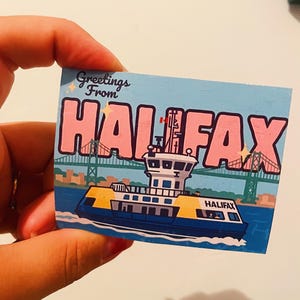 Può includere: Un adesivo rettangolare con il testo Greetings From HALIFAX in lettere rosa. Il design include un traghetto blu e giallo con la scritta HALIFAX sull'acqua, con un ponte verde sullo sfondo sotto un cielo azzurro.