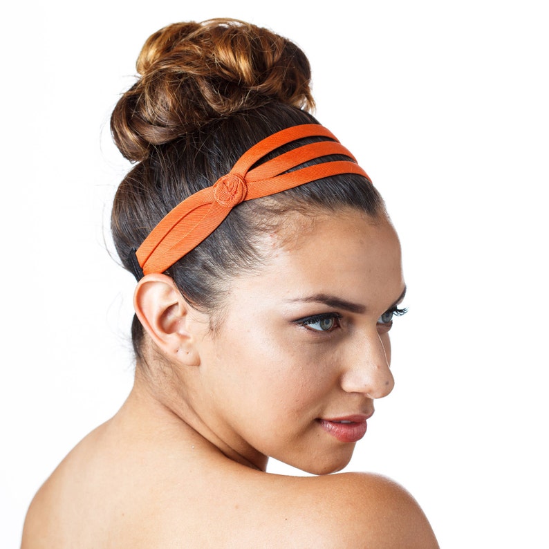 May include: A woman with brown hair styled in a bun wears a bright orange headband with a single button detail. The headband is made of a soft, stretchy fabric.