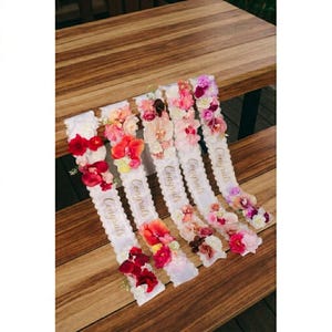 Puede incluir: Seis bandas de satén blanco con arreglos florales y la palabra "Congrats" en escritura dorada. Las flores son en tonos de rojo, rosa y melocotón. Las bandas están dispuestas sobre una superficie de madera.