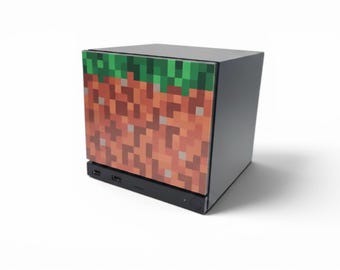 May include: A square-shaped electronic device featuring a pixelated dirt and grass block graphic on its front panel. The unit has a solid dark gray casing, a black base with visible USB ports, and a smooth, flat top surface with a clean, modern design.