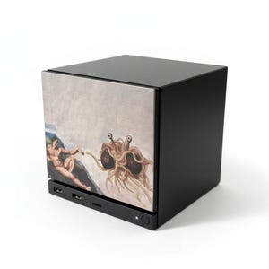 May include: A black cube-shaped electronic device featuring a front panel printed with a parody of The Creation of Adam, depicting a human figure reaching towards a Flying Spaghetti Monster. The base includes two USB ports and a small circular power button.