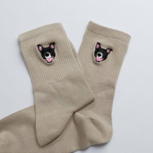May include: A pair of tan socks with a ribbed cuff. Each sock features an embroidered black dog face with pink accents. The socks are made of a soft material and are suitable for everyday wear.