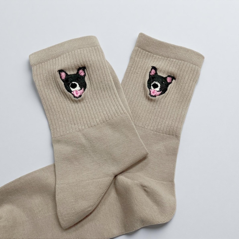 May include: A pair of tan socks with a ribbed cuff. Each sock features an embroidered black dog face with pink accents. The socks are made of a soft material and are suitable for everyday wear.