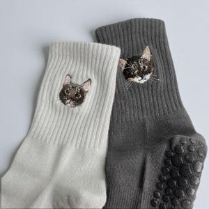 May include: Two pairs of socks, one white and one gray, each embroidered with a cat face. The gray sock has non-slip grips on the sole. The cat embroidery features detailed facial features.