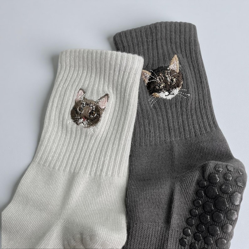 May include: Two pairs of socks, one white and one gray, each embroidered with a cat face. The gray sock has non-slip grips on the sole. The cat embroidery features detailed facial features.