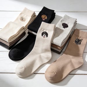 May include: A selection of socks with embroidered animal faces. The socks are in shades of black, beige, and grey. The dog breeds include a golden retriever, a black lab, and a Portuguese water dog. The cat has green eyes.
