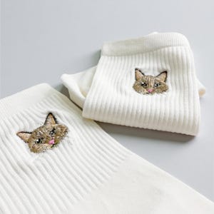 Könnte beinhalten: Cremefarbene gerippte Socken mit gestickten Katzenköpfen. Die Katzenköpfe sind mit braunem, beigem und rosa Garn detailliert, mit ausdrucksstarken Augen und einer kleinen rosa Zunge. Die Socken sind auf hellem Hintergrund gefaltet.