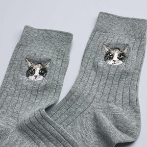 May include: A pair of gray ribbed socks, each featuring an embroidered cat face. The cat's face is detailed with black, white, and brown fur, with green eyes and a pink nose. The socks are made of a soft, comfortable material.