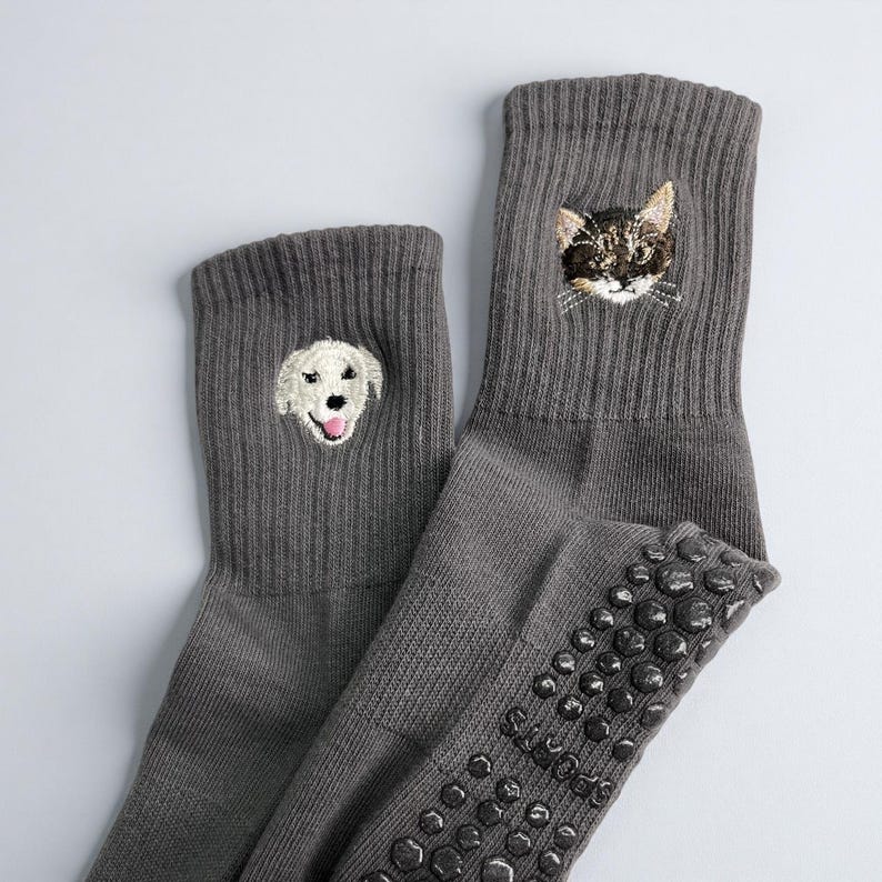 May include: A pair of gray socks with embroidered animal faces. One sock features a dog's face, and the other has a cat's face. The socks have a ribbed texture and non-slip grips on the soles.