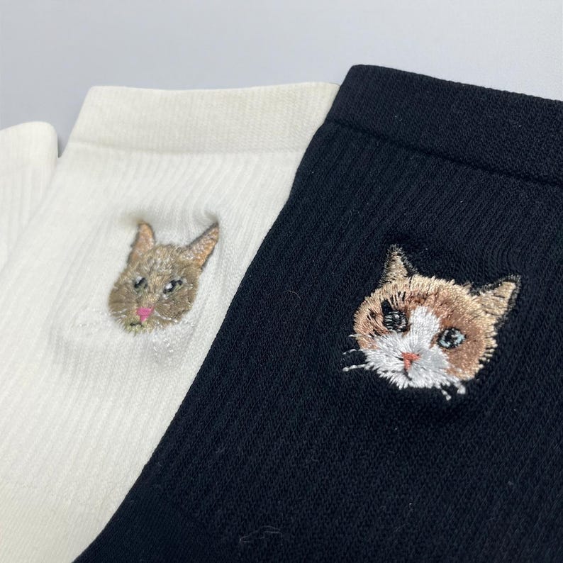 May include: Close-up of two socks, one white and one black, each with an embroidered cat face. The white sock features a gray cat face with a pink nose. The black sock has a brown and white cat face with blue eyes.