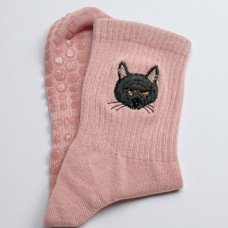 May include: A pair of light pink socks with a black cat face embroidered on the side. The cat has yellow eyes and whiskers. The soles of the socks have small, pink, circular grips.