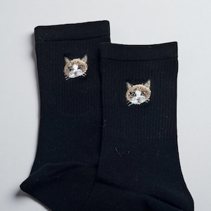 May include: A pair of black socks with an embroidered cat face on each sock. The cat's face is detailed with brown, white, and tan thread. The socks are made of a ribbed knit material.