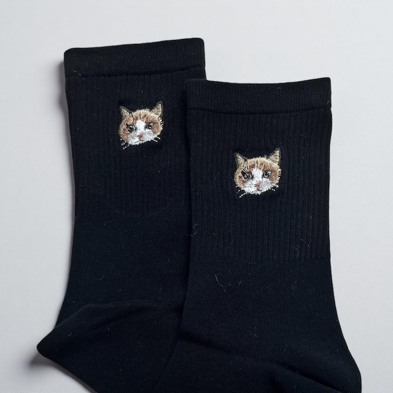 May include: A pair of black socks with an embroidered cat face on each sock. The cat's face is detailed with brown, white, and tan thread. The socks are made of a ribbed knit material.