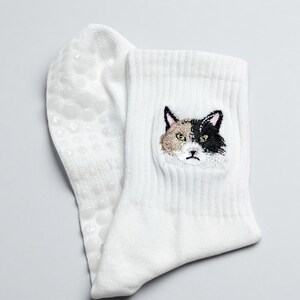 May include: White socks with a cat face embroidered on the front. The cat's face is a mix of black, tan, and white. The bottom of the sock has small, clear, non-slip grips.