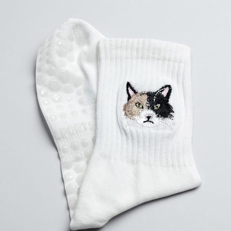 May include: White socks with a cat face embroidered on the front. The cat's face is a mix of black, tan, and white. The bottom of the sock has small, clear, non-slip grips.